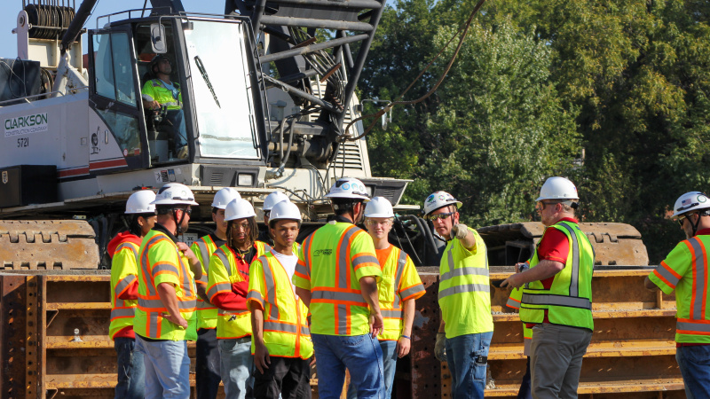 Students in safety gear gather in front of large construction crane.