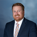 Photo of man in dark-colored jacked with light blue and orange tie