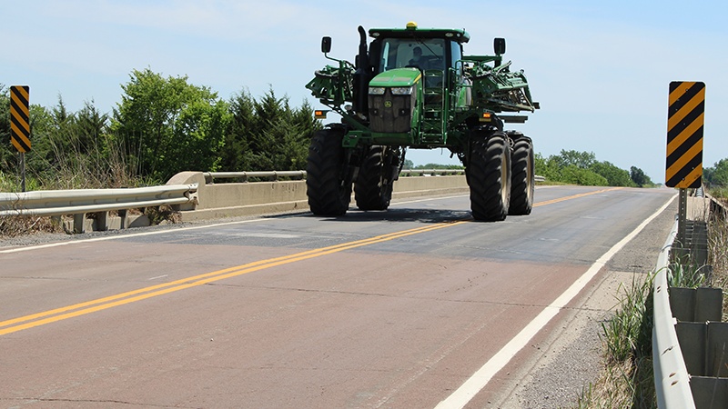 A large piece of green agricultural equipment being driven across the Caldwell County Route 116 bridge over the UP Railroad