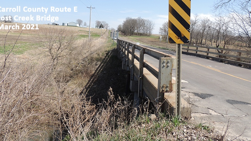 Photo showing the bridge railing, grassy banks and deteriorating surface of the Carroll County Route E Lost Creek Bridge