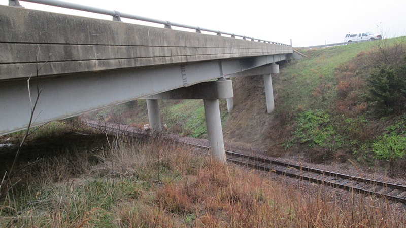 A photo taken from the grassy bank beside the Caldwell County Route 116 bridge over the UP Railroad showing the ednge of the bridge, the supporting structures, and the railroad tracks below