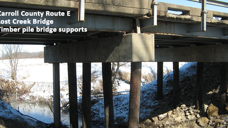 Photo showing the deteriorating concrete on the edge of the bridge's deck and the timber bridge supports out of alignment.
