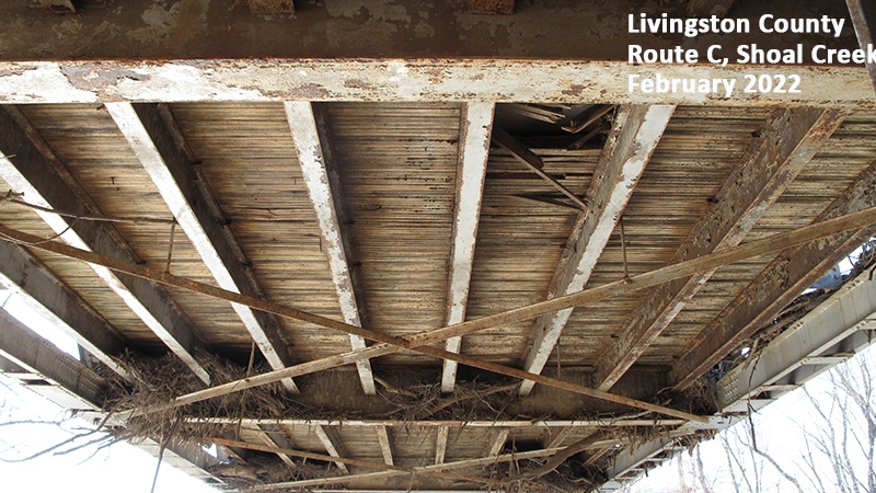 Photo taken directly under the bridge, looking up, showing the wooden timbers - some broken - under the asphalt driving surface of the bridge and debris from flooding caught in the bridge's supporting structures