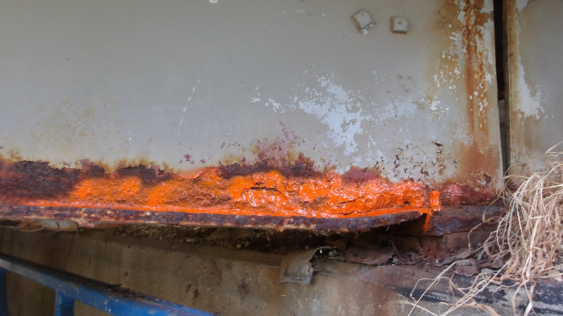 Rust‑damaged metal girder with bright orange paint identifying corrosion along the lower edge, showing structural deterioration and debris.