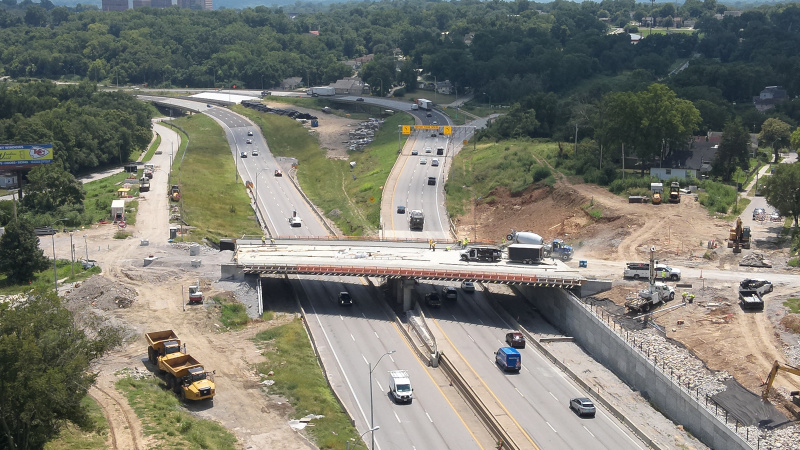 Bridge under construction over an active highway with trees and buildings in the background