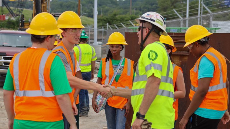 Students in safety gear shake hands with a construction worker